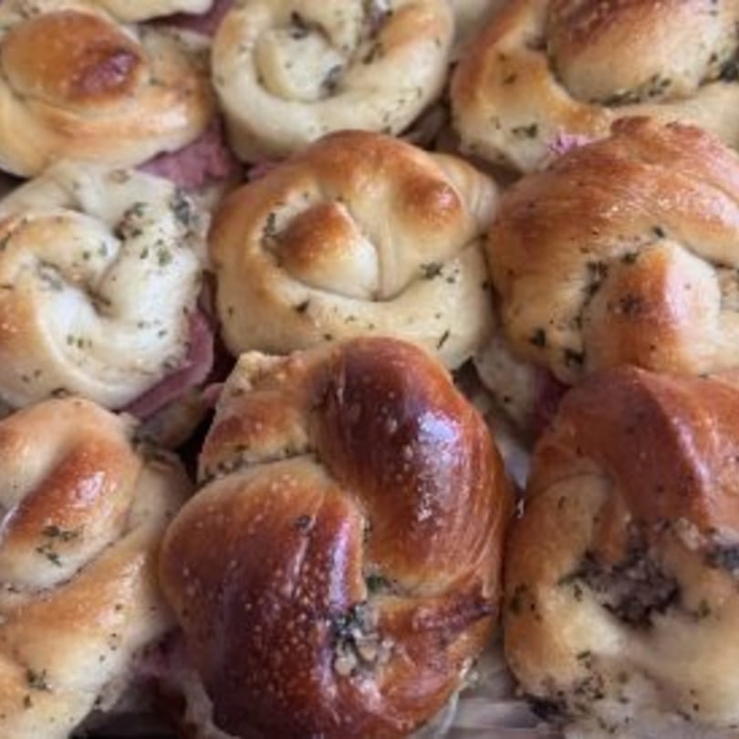 Close-up of several golden-brown, braided garlic knots with herbs, some revealing pink meat filling.