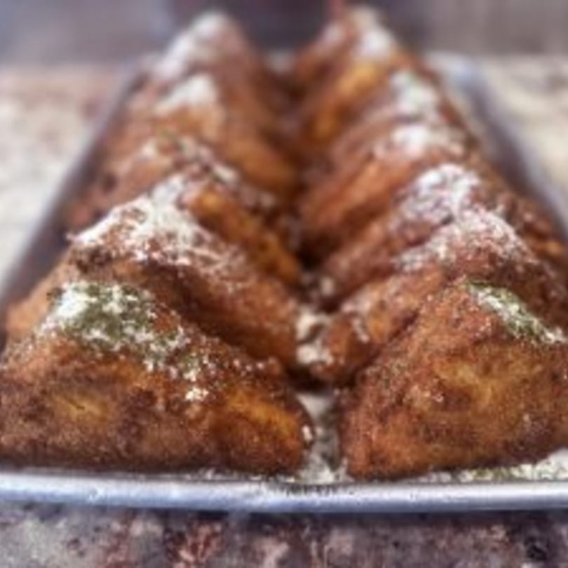 A tray of golden-brown fried dough triangles, dusted with powdered sugar and green herbs.